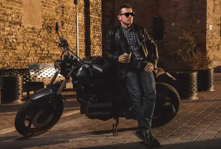 Man wearing a black leather biker jacket standing beside a motorcycle outdoors